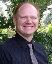 Jon Amon smiling with receding short red hair wearing navy blue button down shirt with gray blue tie and trees in background