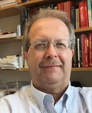 Dr. Ed Brush smiling with short light brown/gray hair and mustache and wearing clear rim glasses and pinstripe white and blue button down shirt with office bookshelves in background