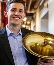 Austin Comerford wearing a black blazer over a blue and white check button down shirt and holding a tuba in his arm