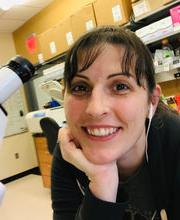 Dr. Joslyn Mills next to microscope in lab smiling with long dark brown hair pulled back in a pony tail with bangs and wearing earphones and a black long sleeve top