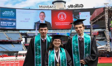 Two men and a woman stand in graduation garb wearing smiles 