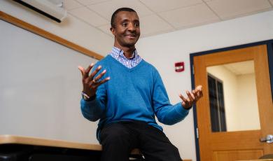 Wayne Magee is sitting on a desk wearing a blue sweater smiling with his hands held up in the air 
