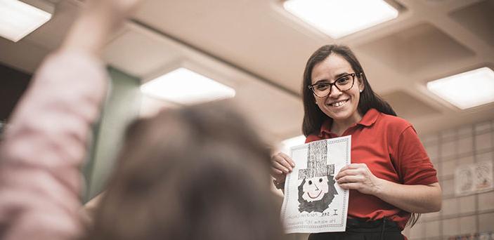 BSU student teacher smiling 和 holding up a childlike drawing of Abraham Lincoln while a student raises her h和